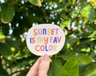 May include: An oval sticker with the text "SUNSET IS my FAV COLOR" in colorful, playful lettering. The sticker is held against a backdrop of green leaves and foliage.