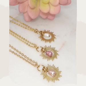 May include: Three gold necklaces with a sunburst pendant and a pearl. The pearls are white, pink, and a light peach color.
