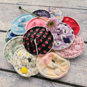 May include: A collection of reusable nursing pads in various floral patterns and colors, including pink, blue, and black. Each pad features a button closure and is arranged around a pink flower with a green stem.