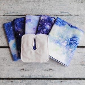 May include: A collection of reusable cloth pads and matching fabric squares. The pads are white, and the fabric squares feature a blue, purple, and white galaxy print. The items are arranged on a light-colored wooden surface.