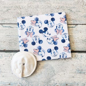 May include: White fabric square patterned with blue and black Mickey Mouse faces and pink bows. A round, white nursing pad with two small buttons is next to the fabric. The items are on a light wood surface.