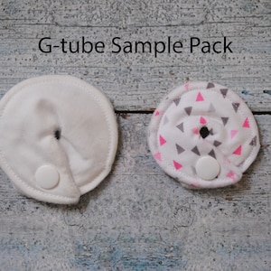 May include: Two round, white fabric G-tube covers with a small hole in the center. One cover is plain white, and the other has a pink and gray triangle pattern. Both covers have a white button on the side.