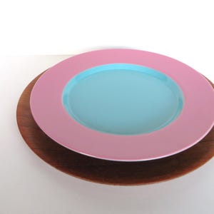May include: A stack of decorative plates. The top plate is light pink with a light blue center. It sits on a dark brown wooden plate. The plates are on a white surface.