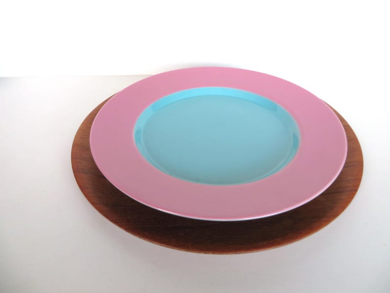 May include: A stack of decorative plates. The top plate is light pink with a light blue center. It sits on a dark brown wooden plate. The plates are on a white surface.
