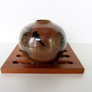May include: A round, brown ceramic vase with black and white glaze. The vase is sitting on a wooden coaster.