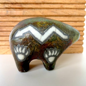 May include: A ceramic bear figurine with a brown, green, and white glaze. The bear has a white zigzag pattern on its back and white paw prints on its sides.