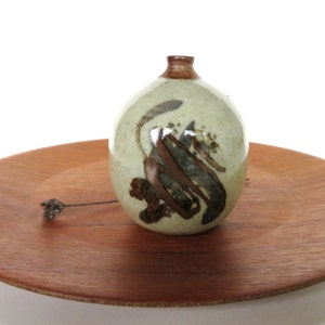 May include: A small, round, ceramic vase with a brown and white glaze. The vase has a brown design on the front and is sitting on a wooden tray.