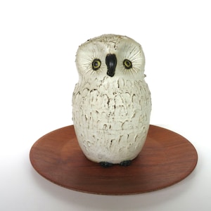 May include: A white owl figurine with textured details, black beak, and yellow eyes, sits on a round, dark brown wooden plate. The owl is a decorative item, likely for home decor.