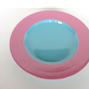 May include: A round, two-toned plate with a pink outer rim and a light blue center. The plate is made of a glossy material and is set against a white background. This decorative plate is suitable for serving food.