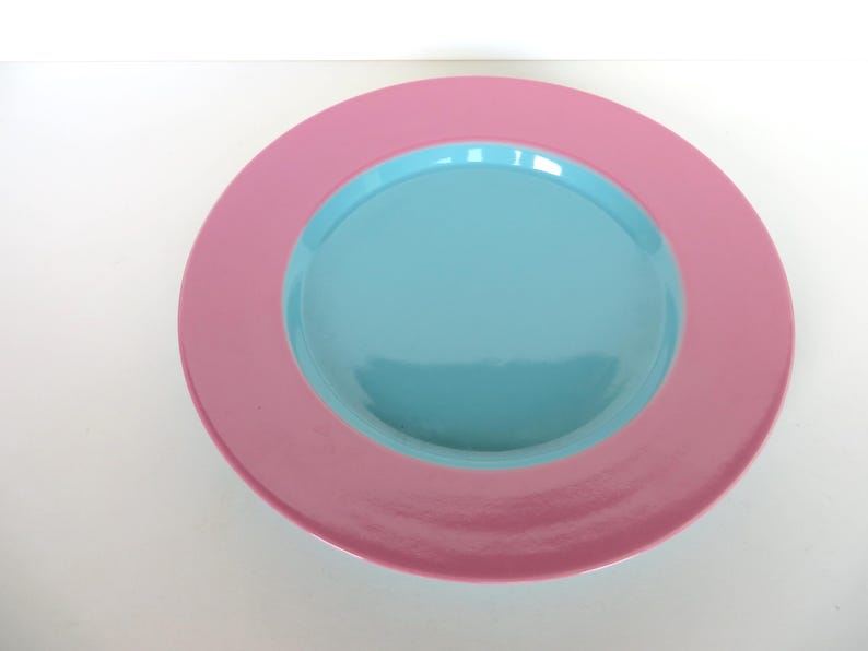 May include: A round, two-toned plate with a pink outer rim and a light blue center. The plate is made of a glossy material and is set against a white background. This decorative plate is suitable for serving food.