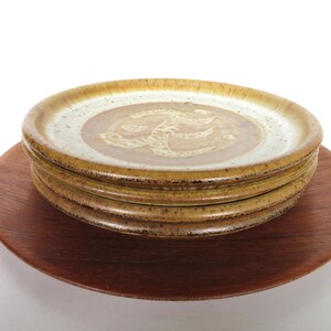May include: A stack of four ceramic plates with a brown and tan color scheme, resting on a wooden charger. The plates have a speckled, textured appearance with a central design. The charger is a dark brown color.