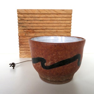May include: A brown ceramic cup with a white interior and a black abstract design. The cup is sitting on a white surface next to a wooden slatted board.