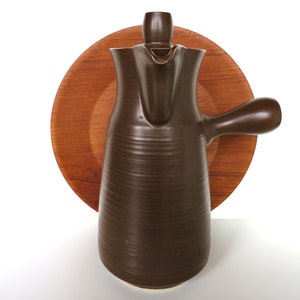 May include: A brown ceramic coffee pot with a wooden base. The pot has a rounded body and a spout on the side. The base is a circular wooden disc.