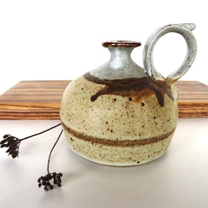 May include: A small, speckled, brown and white ceramic pitcher with a brown glaze and a single handle. The pitcher is sitting on a wooden surface.