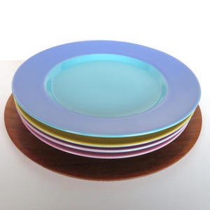 May include: A stack of colorful ceramic plates in shades of blue, turquoise, pink, and yellow, resting on a round wooden charger. The plates have a wide rim and a shallow center. A simple, elegant design for dining.