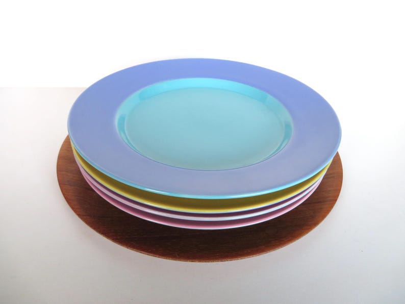May include: A stack of colorful ceramic plates in shades of blue, turquoise, pink, and yellow, resting on a round wooden charger. The plates have a wide rim and a shallow center. A simple, elegant design for dining.