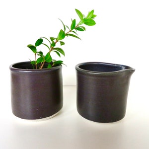 May include: Two dark gray ceramic containers, one with a small green plant and the other a pitcher.