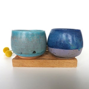 May include: Two ceramic cups, one blue and one teal, with a speckled glaze, sitting on a wooden stand.