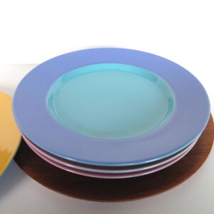 May include: A stack of colorful ceramic plates. The top plate is light blue with a turquoise center. Other plates are pink, white, and yellow. The plates are stacked on a wooden base.