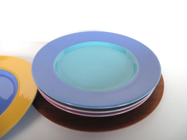 May include: A stack of colorful ceramic plates. The top plate is light blue with a turquoise center. Other plates are pink, white, and yellow. The plates are stacked on a wooden base.