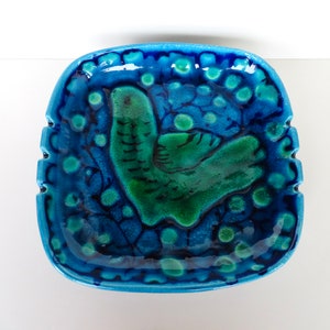 May include: A blue and green ceramic ashtray with a stylized bird design. The ashtray has a square shape with rounded corners and a textured surface.