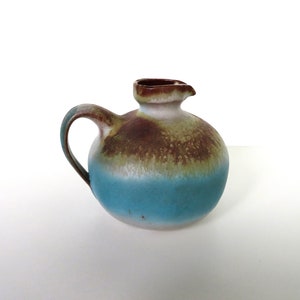 May include: A small, round ceramic pitcher with a handle and spout. The pitcher is painted with a gradient of colors, including blue, green, and brown. The handle is curved, and the spout is small and pointed. The pitcher is likely used for pouring liquids.