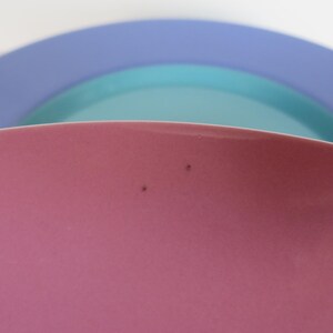 May include: A close-up of three overlapping ceramic plates in shades of pink, teal, and blue. The pink plate is in the foreground, with a slight curve visible. The teal and blue plates are in the background, creating a layered effect.