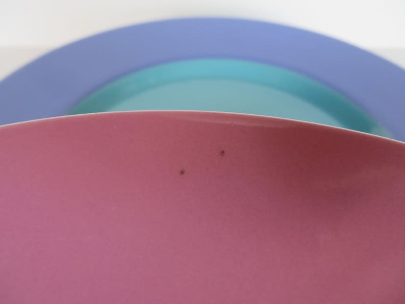 May include: A close-up of three overlapping ceramic plates in shades of pink, teal, and blue. The pink plate is in the foreground, with a slight curve visible. The teal and blue plates are in the background, creating a layered effect.