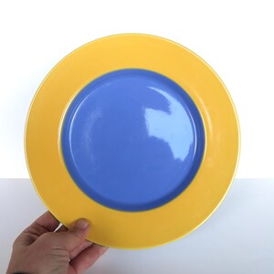 May include: A round plate with a yellow rim and a blue center. The plate is held by a hand against a white background. The plate's design is simple and modern, suitable for everyday use.