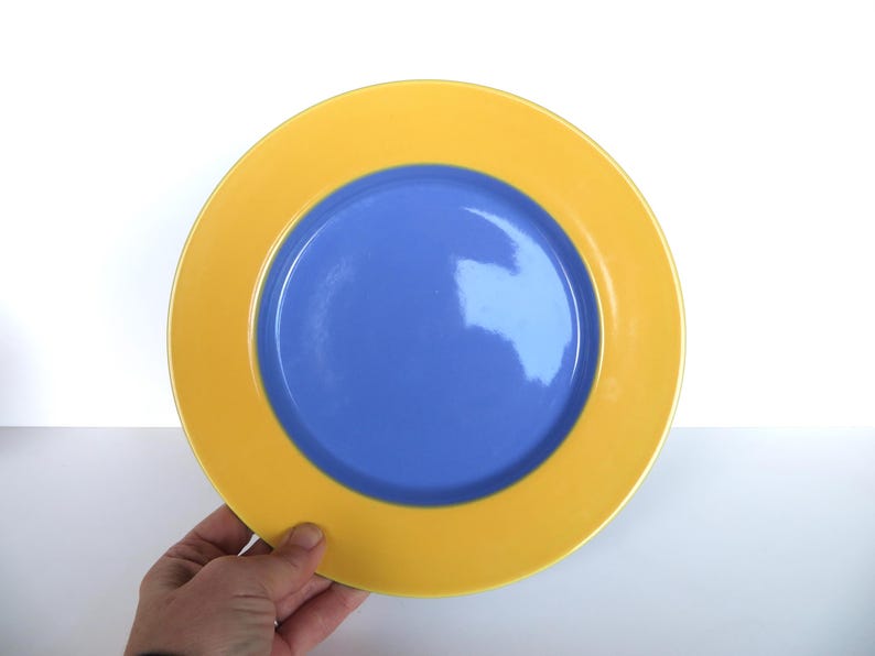 May include: A round plate with a yellow rim and a blue center. The plate is held by a hand against a white background. The plate's design is simple and modern, suitable for everyday use.