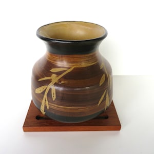 May include: A brown and black ceramic vase with a gold leaf design. The vase is sitting on a wooden coaster.