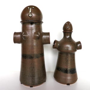 May include: Two brown ceramic vessels with a dark glaze and horizontal black bands. Each vessel features a rounded lid with a simple face, protruding side arms, and a cylindrical body. The taller vessel has a larger head and wider base.
