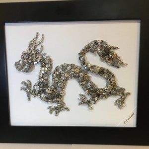 May include: A silver and gold dragon made of buttons and beads is framed in a black frame. The dragon is on a white background. The artist's signature, "P. Castero", is in the lower right corner.