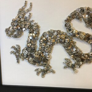 May include: A silver and white dragon made of buttons, beads, and other small metal objects. The dragon is curled up in a classic pose with its head raised and its tail curled around its body.