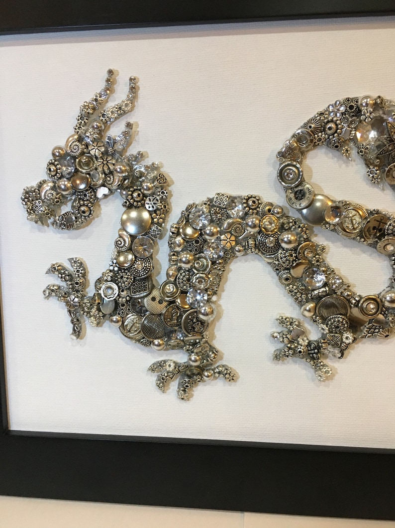 May include: A silver and white dragon made of buttons, beads, and other small metal objects. The dragon is curled up in a classic pose with its head raised and its tail curled around its body.