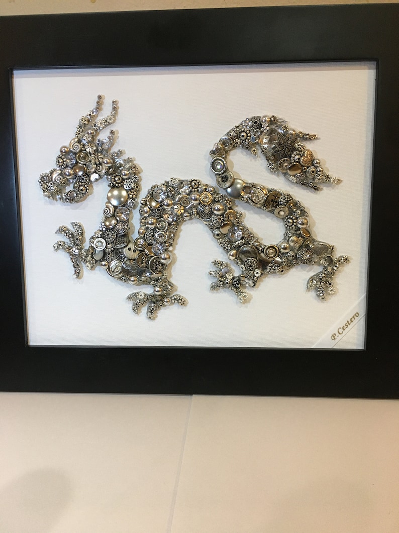 May include: A silver and gold dragon made of buttons and beads is framed in a black frame. The dragon is on a white background. The artist's signature, "P. Castero", is in the lower right corner.