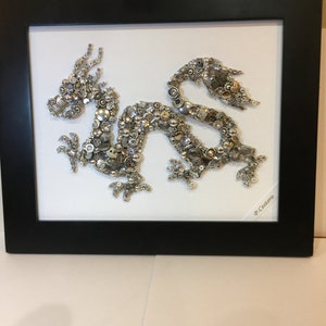 May include: A silver and gold dragon made of buttons is framed in a black frame. The dragon is on a white background and the artist's signature, "P. Castero", is in the bottom right corner.