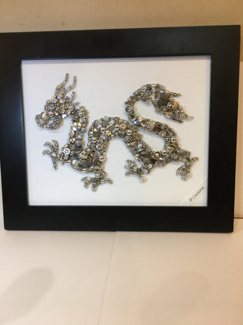 May include: A silver and gold dragon made of buttons is framed in a black frame. The dragon is on a white background and the artist's signature, "P. Castero", is in the bottom right corner.