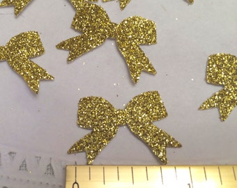 May include: Gold glitter bows, approximately 1 inch in size, scattered on a white surface with a ruler for scale.
