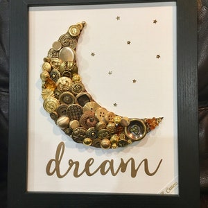 May include: A framed artwork featuring a crescent moon made of gold buttons and a gold glitter background with the word "dream" written in gold script.
