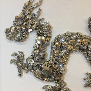 May include: A silver and gold dragon made of buttons and beads on a white canvas background.