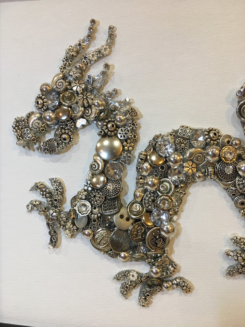 May include: A silver and gold dragon made of buttons and beads on a white canvas background.