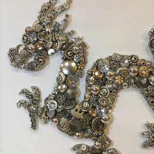 May include: A silver and gold dragon made of buttons, beads, and other small metal objects. The dragon is on a white canvas background.