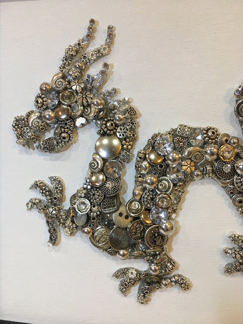 May include: A silver and gold dragon made of buttons, beads, and other small metal objects. The dragon is on a white canvas background.