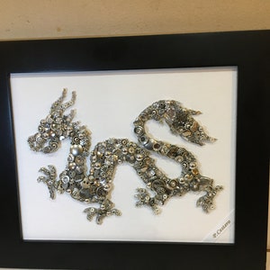 May include: A silver and gold dragon made of buttons and other small objects is framed in a black frame. The dragon is on a white background. The artist's signature, "P. Castero", is in the bottom right corner.