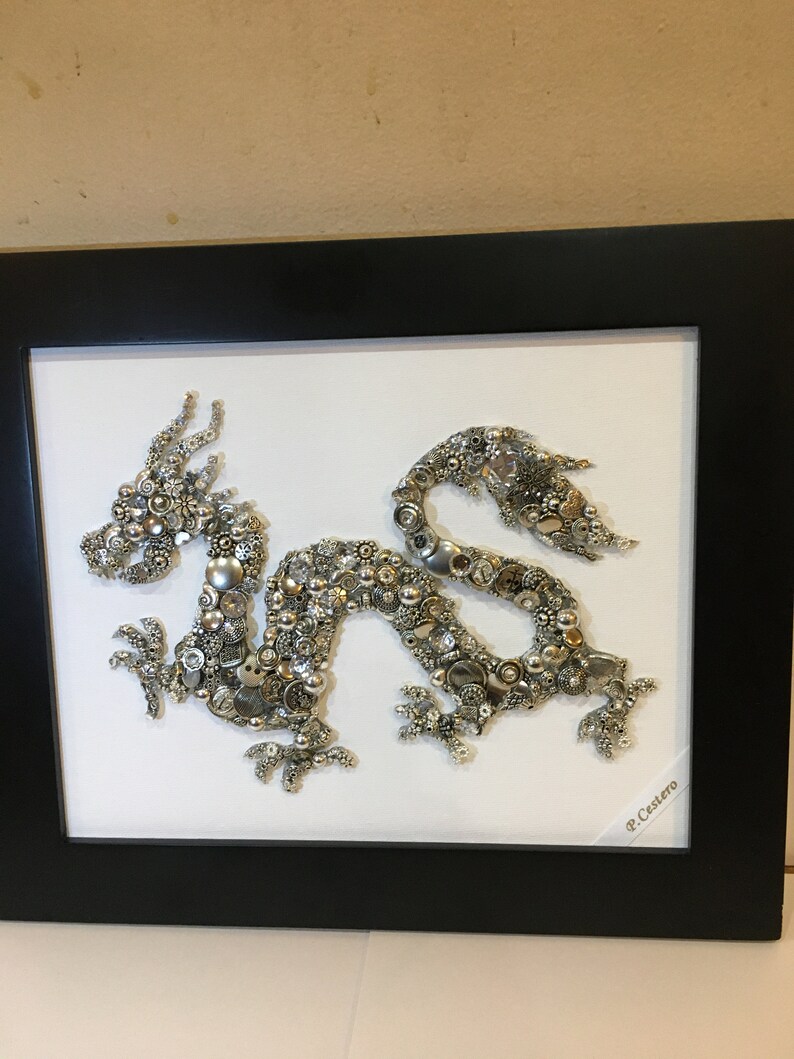 May include: A silver and gold dragon made of buttons and other small objects is framed in a black frame. The dragon is on a white background. The artist's signature, "P. Castero", is in the bottom right corner.