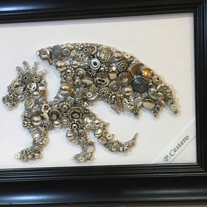 May include: A silver and gold dragon made of buttons and other small objects is framed in a black frame. The artist's signature, "P. Cestero", is visible in the lower right corner.