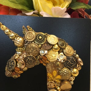 May include: A handcrafted unicorn head silhouette, created from an assortment of vintage gold buttons and embellishments, is displayed against a black background. The artwork is accented with yellow and orange floral elements.
