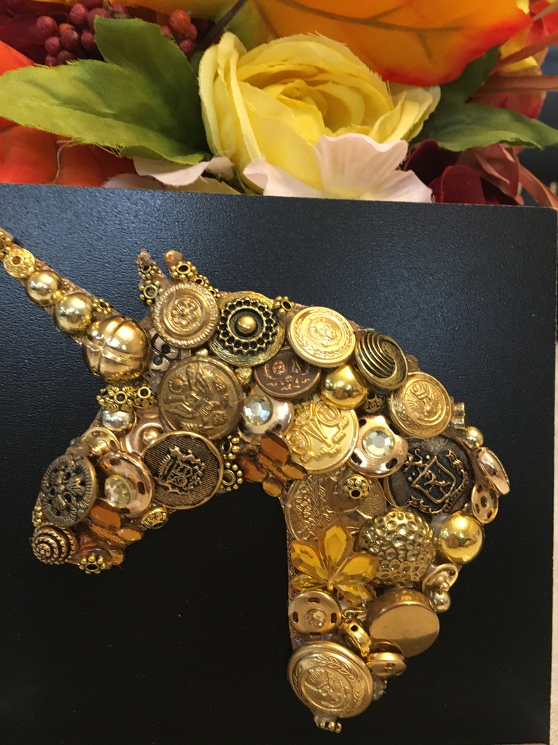 May include: A handcrafted unicorn head silhouette, created from an assortment of vintage gold buttons and embellishments, is displayed against a black background. The artwork is accented with yellow and orange floral elements.