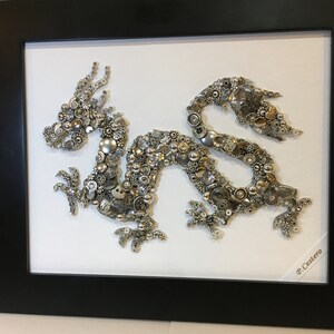 May include: A framed artwork depicting a silver dragon made of buttons and beads. The dragon is set against a white background and the frame is black. The artist's signature, "P. Cestero", is visible in the bottom right corner.